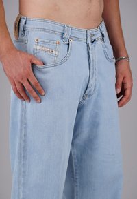 Man wearing light blue jeans with hand in pocket, showing exposed midriff and a silver bracelet on right wrist.