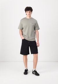 Lys grå bomulds t-shirt med logo på brystet, parret med sorte denim shorts. Model iført sorte sneakers med hvide accenter.