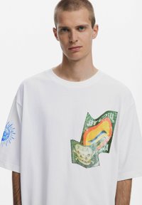 Balta per didelė marškinė su spalvingais grafikos spaudiniais ir mėlyna dekoracija kairiajame rankove. Minkšta medžiaga, plačiojo silueto.