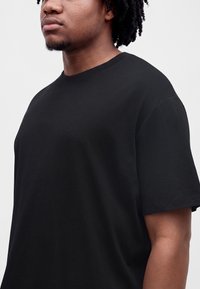 Junge Person mit dunklem lockigem Haar, die ein schlichtes schwarzes T-Shirt mit kurzen Ärmeln vor einem hellgrauen Hintergrund trägt.