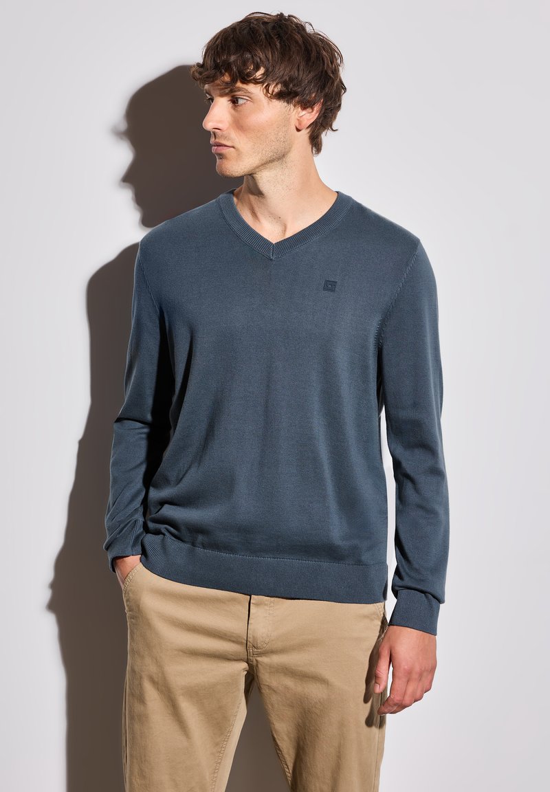 Pull bleu en V en matière douce et à maille fine, avec des manches longues et un petit logo sur la poitrine, associé à un pantalon khaki.