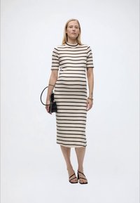 Έγκυος γυναίκα που φοράει μπεζ και μαύρο ριγέ φόρεμα μέχρι το γόνατο, κρατώντας μια μαύρη τσάντα, στέκεται μπροστά σε ένα λευκό, απλό φόντο.