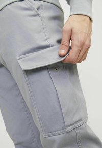 Hand hebt den Deckel der hellgrauen Cargo-Tasche an der passenden Hose an und zeigt den Metall-Druckknopf sowie die Nähdetails.