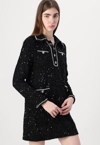 Vestido preto texturizado com elementos brilhantes, decote com gola, bolsos frontais com botões decorativos e detalhes de bainha contrastantes.