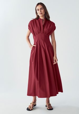 Bordeauxfarbene Maxikleid aus glattem Stoff, mit hohem Kragen, Puffärmeln und Seitentaschen, sowie einer fließenden A-Linie-Silhouette.