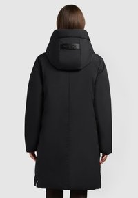 Černý prošívaný kabát s vysokým límcem, s logovou nášivkou na zádech, dlouhými rukávy a detaily se zipy po stranách. Hladká textura látky.