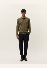Olijfgroene polo met lange mouwen en drie knopen, gecombineerd met donkere marineblauwe pantalon en zwarte schoenen, afgerond met een zwarte baseballcap.