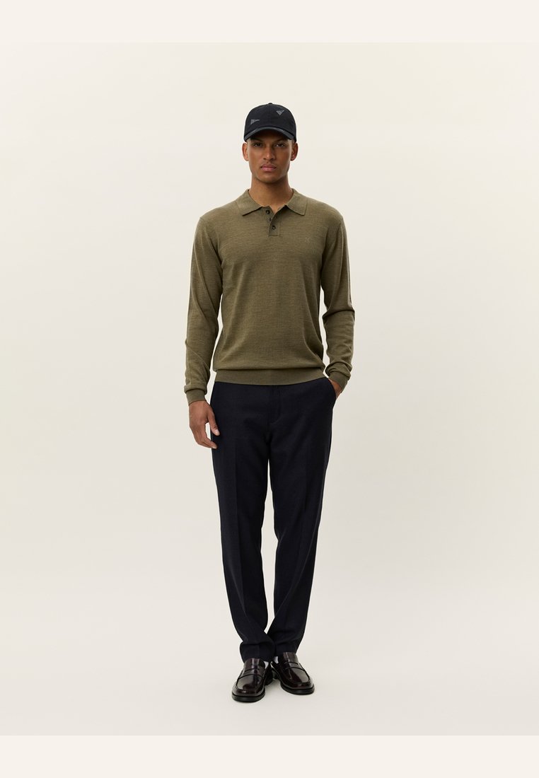 Olijfgroene polo met lange mouwen en drie knopen, gecombineerd met donkere marineblauwe pantalon en zwarte schoenen, afgerond met een zwarte baseballcap.