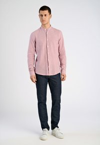 Camicia button-up rosa chiaro con colletto a band, maniche lunghe e taschino sul petto, abbinata a jeans blu scuro e sneakers bianche.
