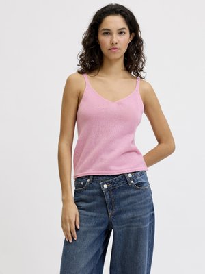 Jeune femme aux cheveux bouclés et foncés portant un haut en maille rose sans manches et un jean bleu à jambes larges sur un fond clair uni.