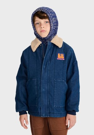 Enfant portant une veste matelassée bleue avec un col en fourrure et une capuche en tricot bleue, debout devant un fond clair uni.