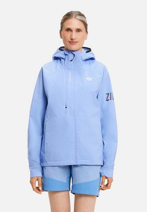 Frau trägt eine hellblaue Kapuzenjacke und passende zweifarbige blaue Shorts, steht vor einem schlichten weißen Hintergrund.