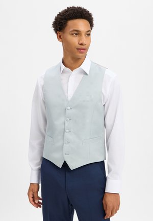 Junger Mann trägt ein weißes Hemd, eine hellblaue gemusterte Weste und dunkelblaue Hose, steht und schaut leicht zur Seite.