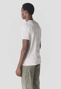 Jeune homme aux cheveux courts et bouclés portant un t-shirt blanc texturé à manches courtes et un pantalon cargo vert olive, vu de profil.