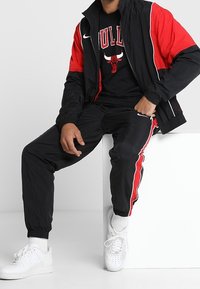 Černá a červená windbreaker bunda Nike v kombinaci s černými tepláky s červenými bočními pruhy, bílými teniskami a černým tričkem s grafikou Bulls.
