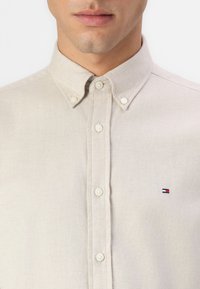 Cămașă bej deschis, cu nasturi, fabricată din material de in; prezintă un guler ascuțit, nasturi albi și un mic logo brodat pe pieptul stâng.