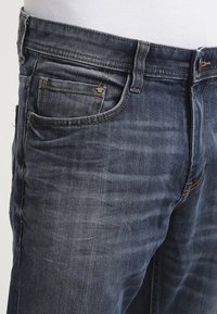 Jeans de mezclilla azul oscuro con un lavado desgastado, que presentan un diseño clásico de cinco bolsillos y costuras visibles. Cierre con botón y cremallera.