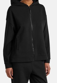Sudadera con cremallera en negro, hecha de una tela suave, que cuenta con una capucha grande, bolsillos frontales y un corte relajado con puños y dobladillo de canalé.