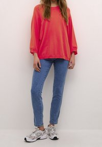 Korallenfarbener, übergroßer Pullover mit langen Ärmeln, kombiniert mit hellblauen Skinny-Jeans und grauen Sportschuhen. Hat eine lässige Passform und eine weiche Textur.