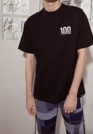 Sort t-shirt med korte ærmer, der har et hvidt tryk med teksten "100 Dickies" på brystet, kombineret med patchwork denimjeans.