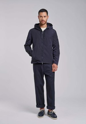 Man staat en draagt een donker capuchonjack, donkere broek met opgerolde pijpen, en marineblauwe bootschoenen met witte veters op een lichtgrijze achtergrond.