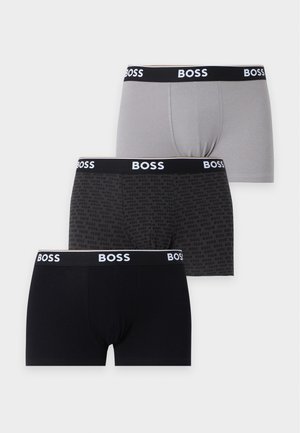Mladý muž s tmavými vlasy, oblečený v šedých boxerkách s černým elastickým páskem BOSS, stojí naboso na jednobarevném bílém pozadí.