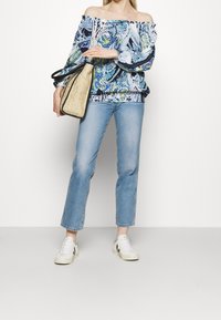Blusa off-shoulder con un motivo floreale blu e verde, abbinata a jeans di denim chiaro e sneakers bianche, mentre tiene una borsa in oro zigrinato.