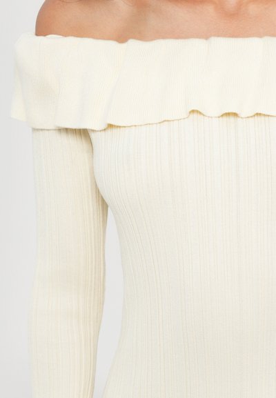 Off-the-shoulder top in light cream ribbed fabric, featuring a ruffled trim and long sleeves, showcasing a fitted silhouette.