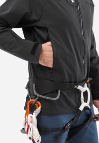 Person wearing a black jacket with hand in zippered pocket and a climbing harness holding carabiners and safety ropes.
