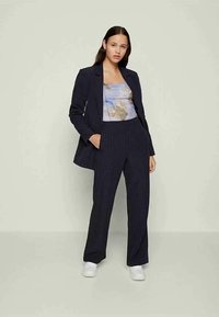 Mulher de pé com as mãos nos bolsos, vestindo um fato azul com riscas, uma blusa estampada e tênis brancos, contra um fundo claro e simples.