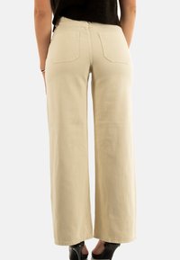 Mujer con pantalones de pierna ancha beige y blusa negra, de pie con los pies en zapatos de tacón negros, vista desde atrás.