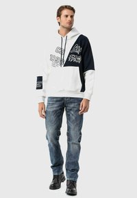 Kapuzenpullover mit navyblauen und weißen Segmenten, ausgestattet mit grafischen Drucken, einer Fronttasche und gerippten Bündchen. Kombiniert mit blauen Jeans und schwarzen Stiefeln.