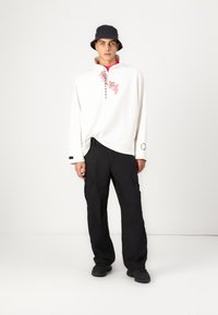 White long-sleeve shirt with pink floral design and black lettering, paired with black cargo pants and a navy bucket hat. Black shoes.