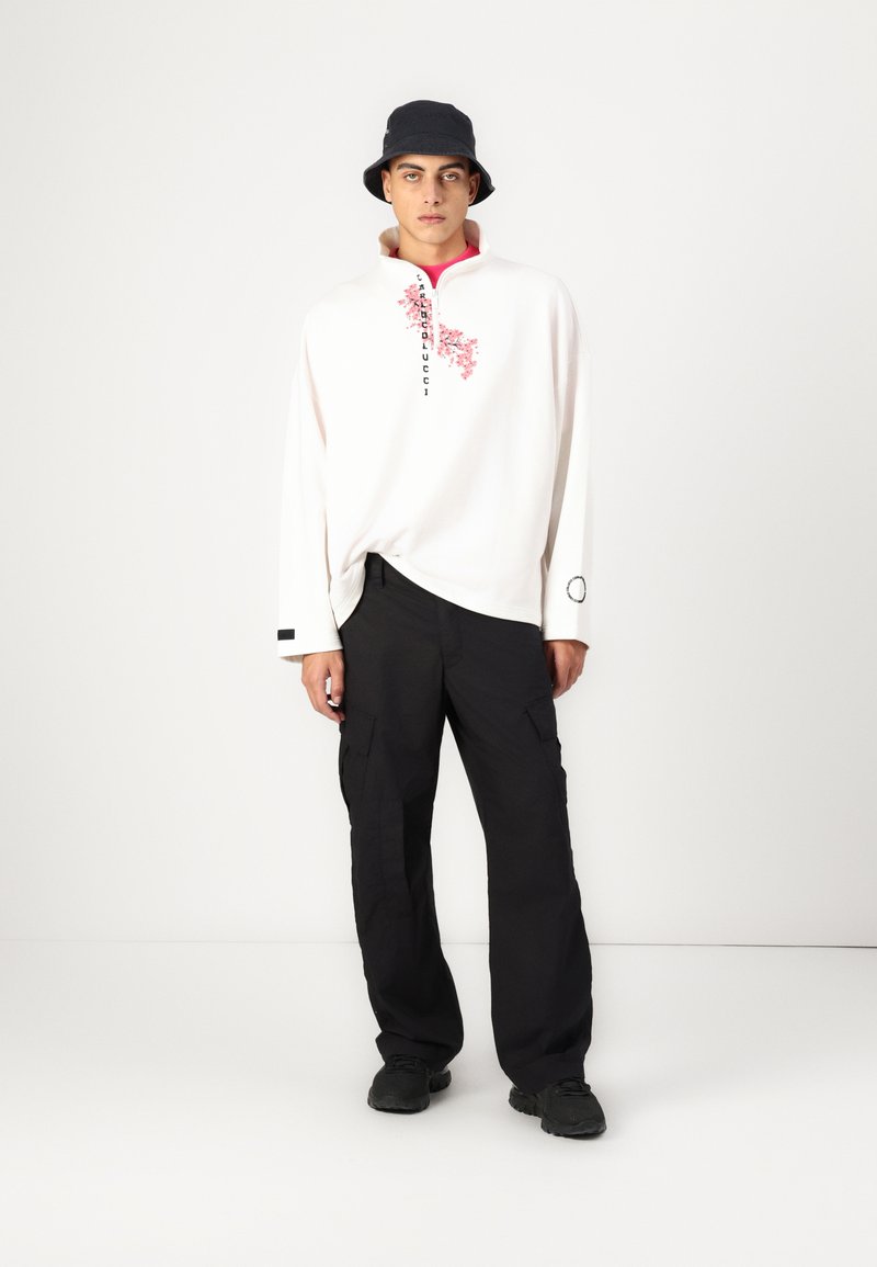 Biała koszula z długim rękawem w różowy kwiatowy wzór, z czarnymi napisami, w połączeniu z czarnymi spodniami cargo i granatowym kapeluszem typu bucket. Czarne buty.