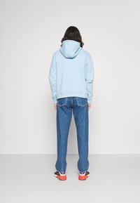 Light blue hoodie with a front pocket, paired with loose blue denim jeans and black sneakers with red accents. Visible stitching and texture details.