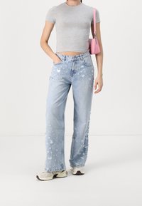 Jeans de denim desgastados de color azul claro con un ajuste relajado, que presentan grandes desgastes y dobladillos deshilachados. Combinados con una camiseta ajustada gris y zapatillas.