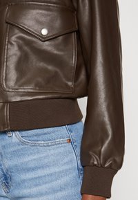 Giubbotto bomber in pelle marrone con polsini e vita a coste, dotato di una tasca a scatto sul petto. Abbinato a jeans in denim blu.