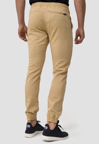 Lysbrune joggebukser med elastisk talje, sidelommer med lynlås og tapered ben. Båret med mørke sneakers, der har tekstureret materiale.