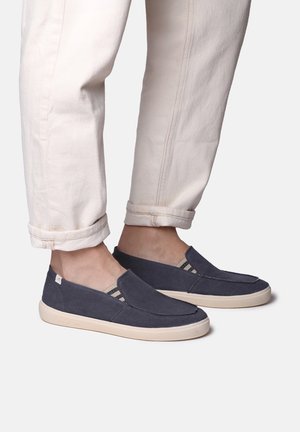 Persona che indossa pantaloni beige chiaro risvoltati e scarpe slip-on blu navy con suole color crema su sfondo bianco.