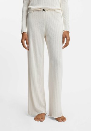 Persona che indossa pantaloni da salotto color crema con maglia a coste e un fiocco nero in vita, abbinati a una maglia a maniche lunghe dello stesso colore, a piedi nudi su sfondo bianco.