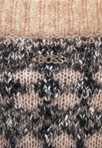 Écharpe en tricot avec des motifs texturés beiges et noirs, ornée d'un petit logo "BOSS" en argent sur le bas.