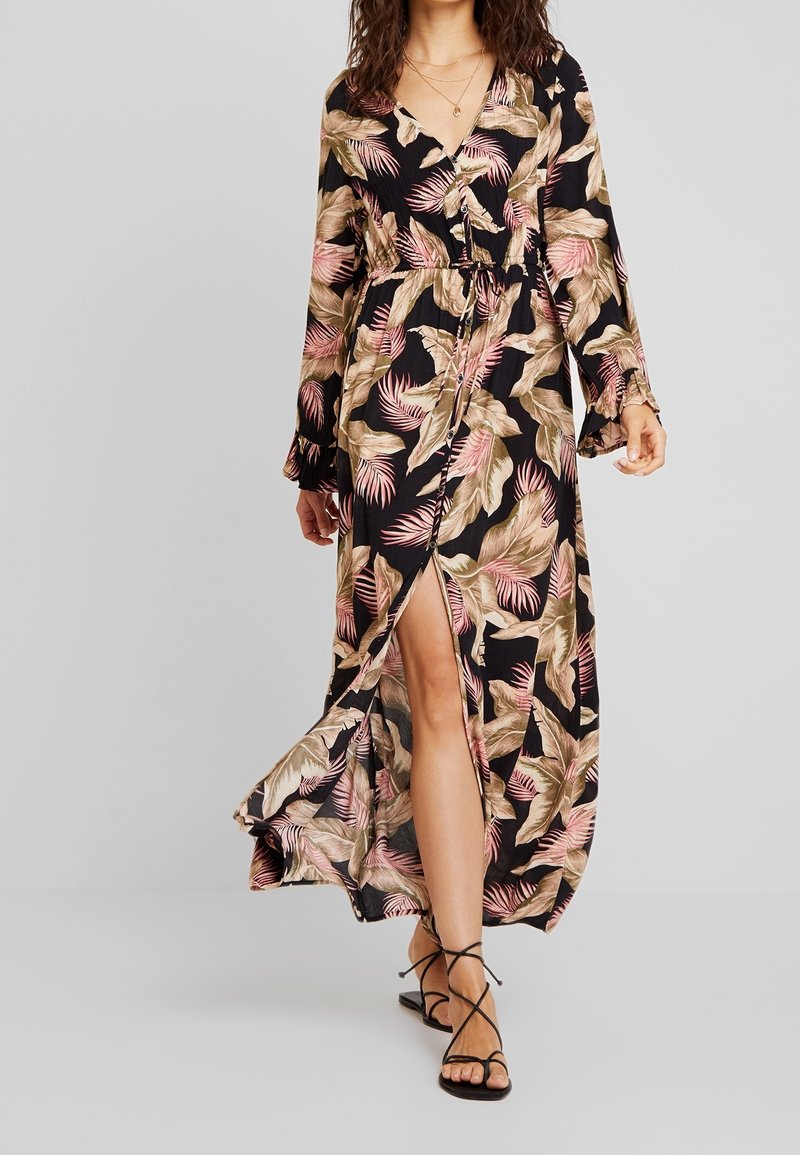 Femme portant une longue robe noire avec des motifs de feuilles roses et beiges, une fente devant, et des sandales noires à brides, debout sur un fond gris.