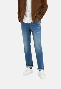 Jeans din denim albastru deschis cu croială dreaptă, cu manșete răsucite, asortate cu o cămașă albă cu model și o jachetă din catifea călcată de culoare maro, purtate cu adidași albi.