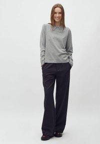 Fiatal nő mosolyog, kezét zsebében tartja, szürke hosszú ujjú pulóvert visel díszített gallérral, sötét színű, bő szárú nadrágot és sötét cipőt.