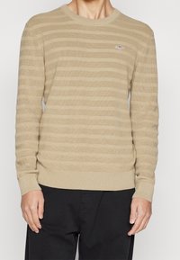 Beige Strickpullover mit abwechselnd hellen und dunklen Streifen, geripptem Halsausschnitt und Bündchen, versehen mit einem kleinen Logo auf der Brust.