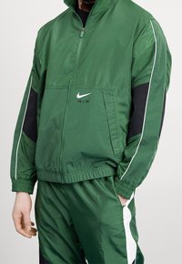 Giacca verde con zip realizzata in materiale leggero, con dettagli neri e piping bianco. Presenta il logo Nike e polsini elastici.