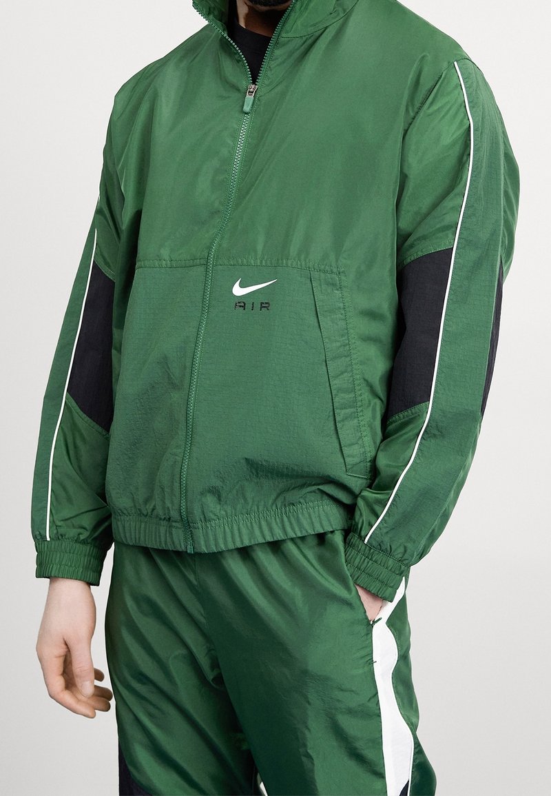 Giacca verde con zip realizzata in materiale leggero, con dettagli neri e piping bianco. Presenta il logo Nike e polsini elastici.