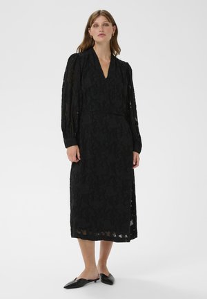 Schwarzes Kleid mit floralem Spitzenmuster, tiefem V-Ausschnitt, langen Ärmeln und fließender Silhouette, kombiniert mit schwarzen, spitzen Ballerinas.