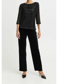 Pantalons en velours noir associés à un haut noir texturé avec des manches 3/4 et un motif subtil. Des escarpins noirs fins complètent l'ensemble.