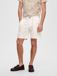 Shorts en lin blancs avec une taille élastique et un cordon de serrage ; associés à une chemise beige à manches courtes avec un motif de feuilles ; chaussures marron foncé.